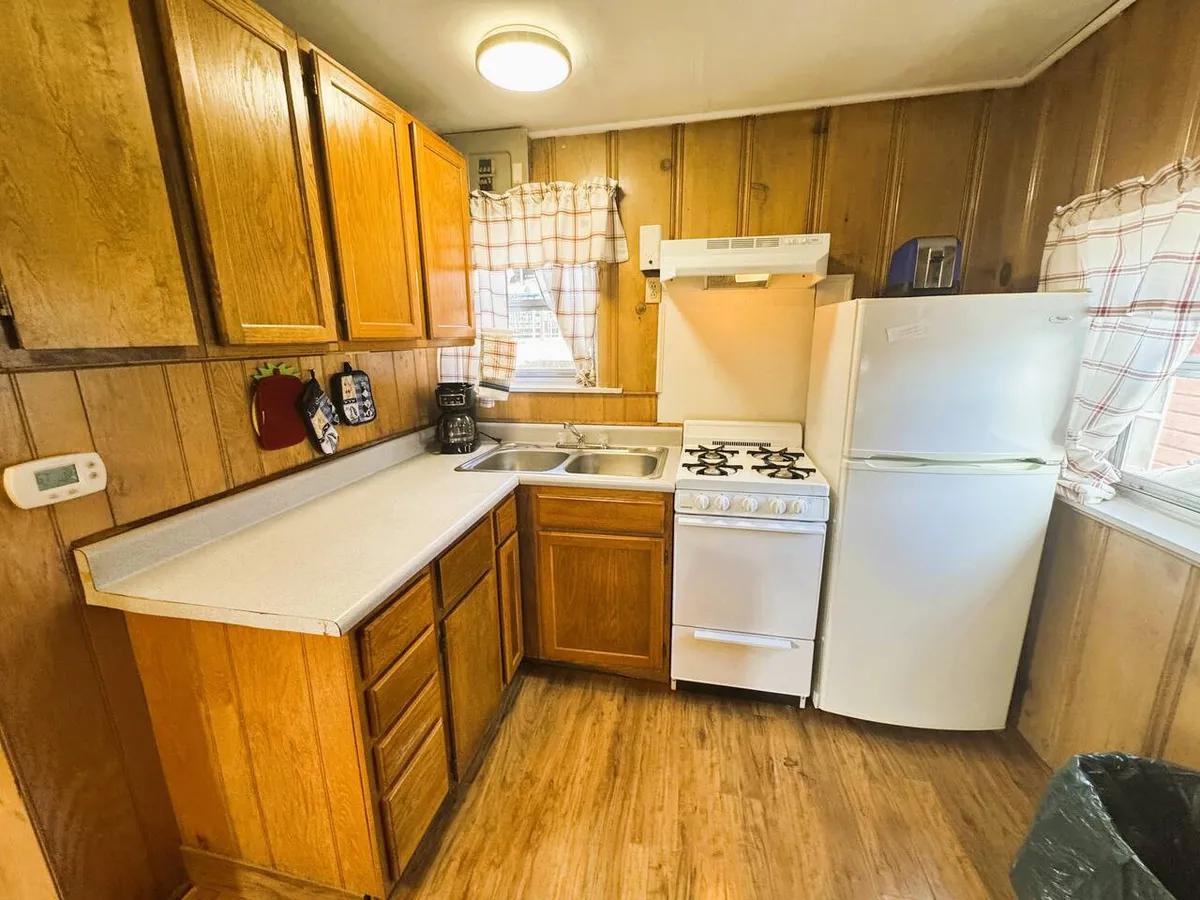 A cozy kitchenette with wooden cabinets, white appliances including a stove and refrigerator, a kitchen sink, and a window with curtains above. The countertop is spacious enough for meal prep, giving a homey, functional feel to the space.