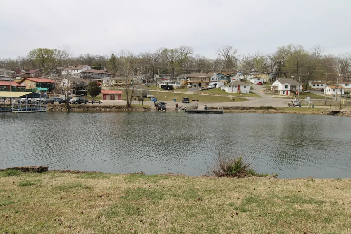 A wide view of calm lake waters with boats and houses along the shoreline. Green grass in the foreground hints at easy lakeside access, and the overall scene exudes peacefulness—ideal for fishing, boating, or spending time by the water.