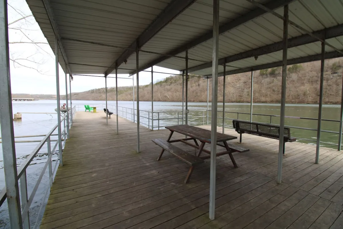 A serene lakeside scene featuring a wooden dock with a picnic table and green chairs, set against a backdrop of calm water and rolling hills. The area is ideal for relaxing, enjoying meals outdoors, and taking in the peaceful surroundings.
