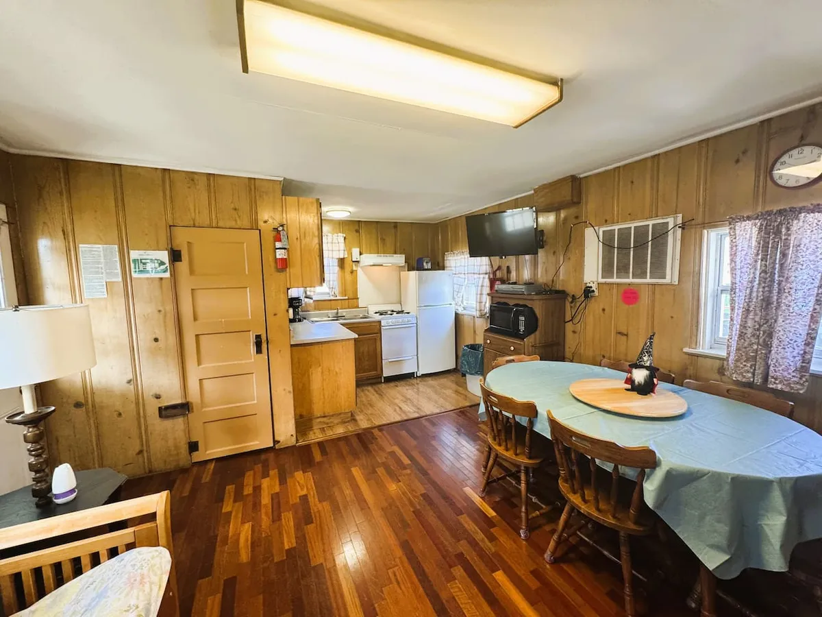 An inviting interior scene featuring a dining table set against wood paneling, with kitchen appliances and a warm lamp in view. The setting suggests family meals, game nights, or relaxed gatherings in a homey environment.