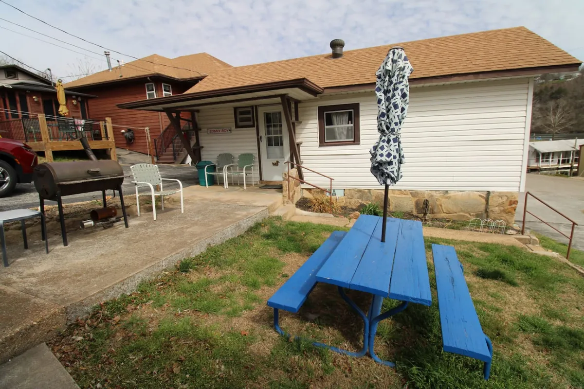 A grassy outdoor area with a blue picnic table under an umbrella, a BBQ grill nearby, and white chairs set up for relaxed outdoor dining. The setting is perfect for family barbecues and enjoying meals al fresco.