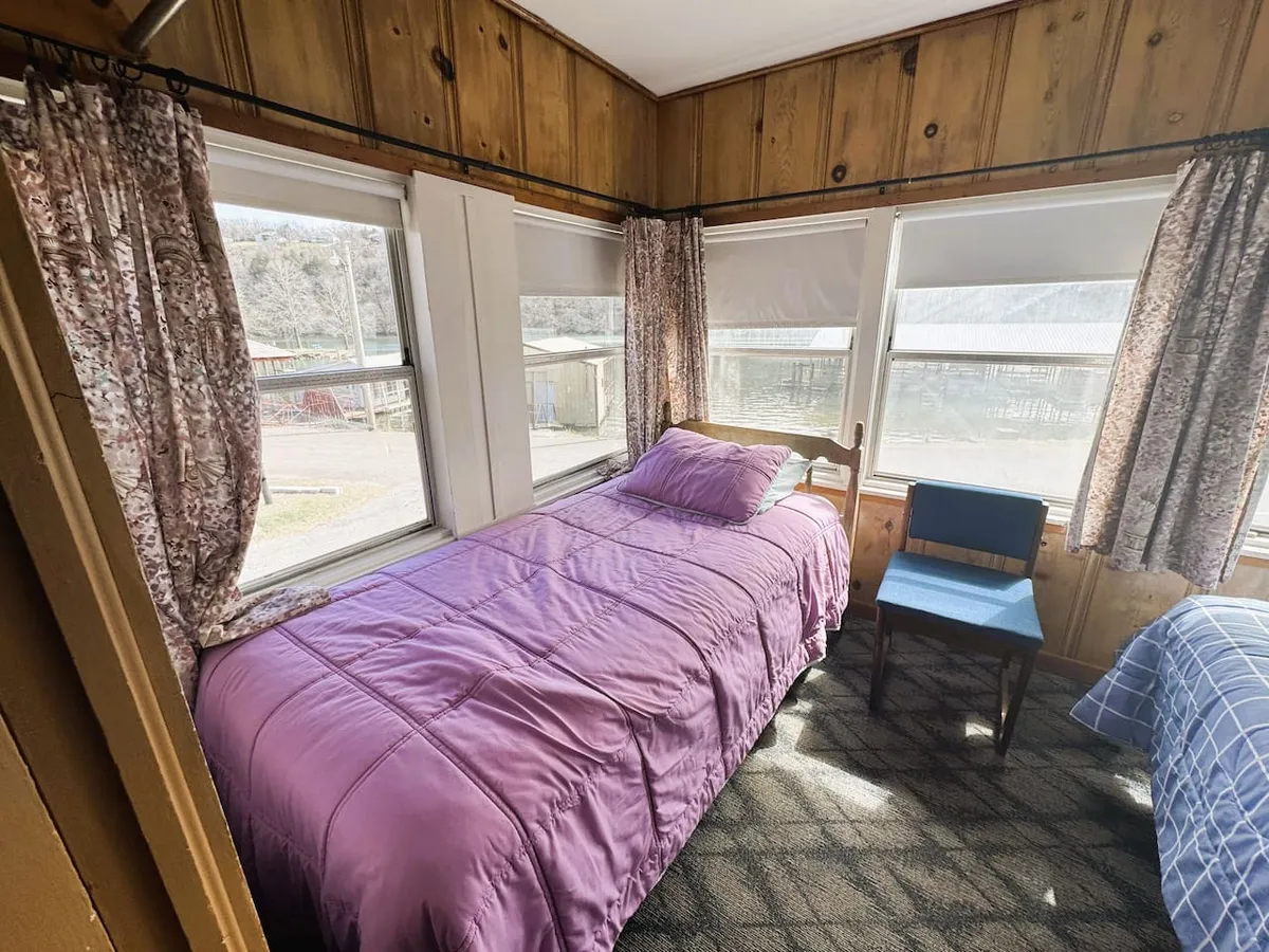 A serene bedroom scene featuring a plush bed with a purple bedspread, a blue chair near the window, and soft curtains that gently filter natural light. The space feels peaceful and private, ideal for rest and relaxation.