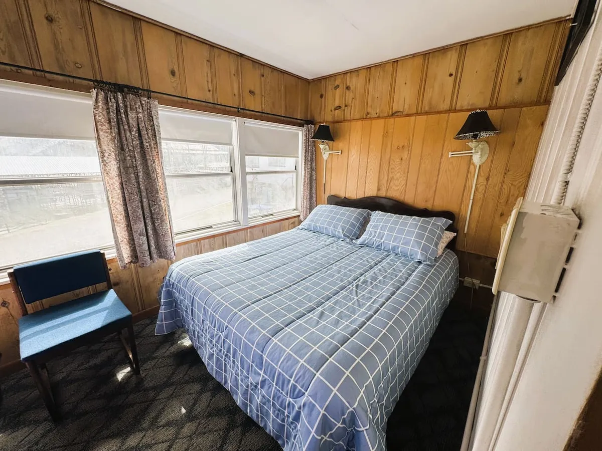 A cozy sleeping and living area with a plush bed, inviting chair, lamps, and curtains that frame large windows letting in an abundance of natural light. The warm tones and soft textures create an atmosphere perfect for relaxing or enjoying a quiet morning with a book.