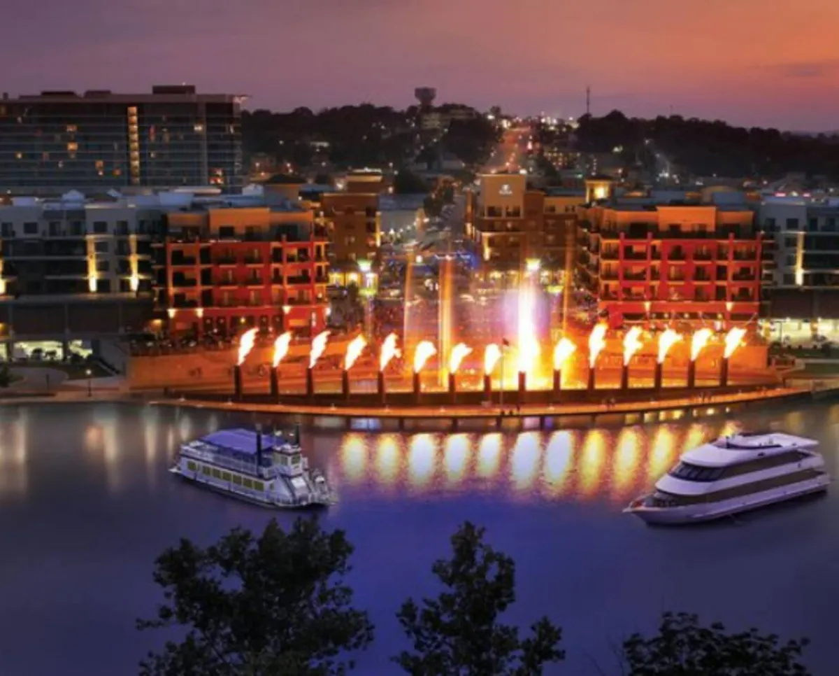 A vibrant waterfront scene featuring a fountain with fire features, boats, and buildings lit up for the evening. People are likely enjoying dining, boat rides, and the lively local atmosphere along the water.