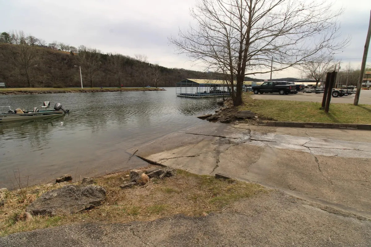 A waterfront scene featuring a boat ramp, a docked boat, trees, and a parking area adjacent to the lake. The area looks ready for boat launching, with clear access from the shore.